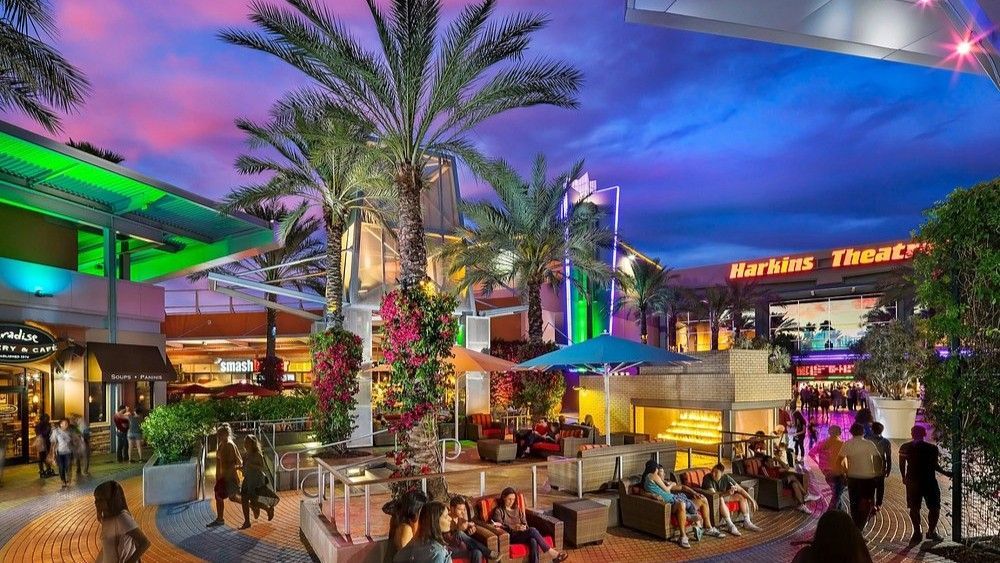 Outdoor mall scene with people, restaurants, and a theater, lit up at dusk with colorful lights.