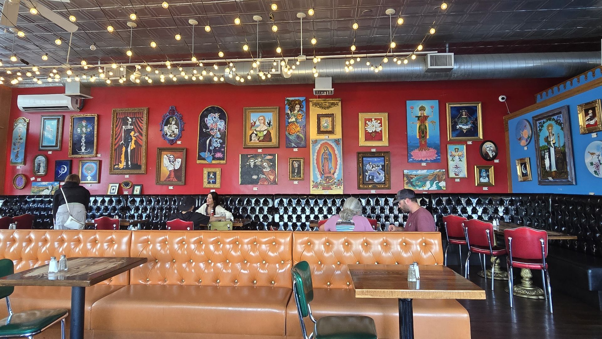 Interior restaurant with red wall, art, booths, tables, and string lights. People are seated.