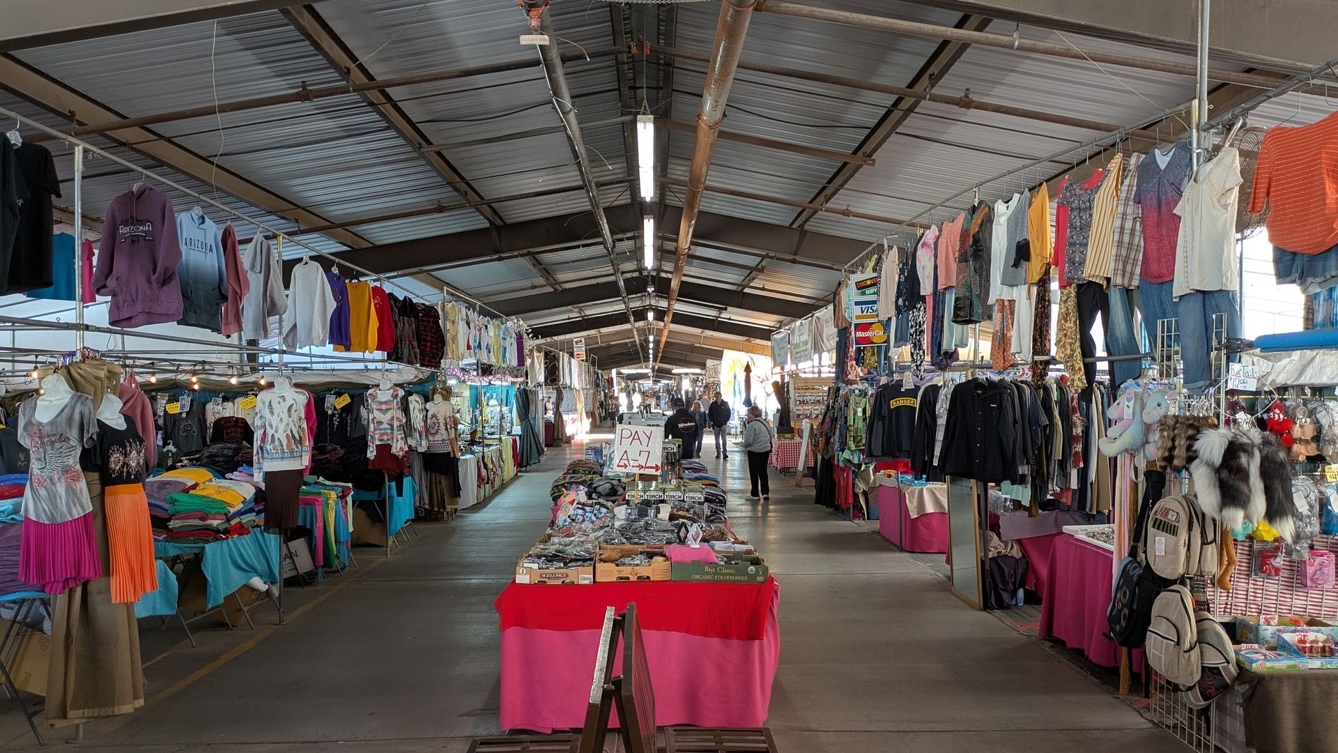 Inside view of a covered market with clothing on display, featuring long aisles, pink and red tables.