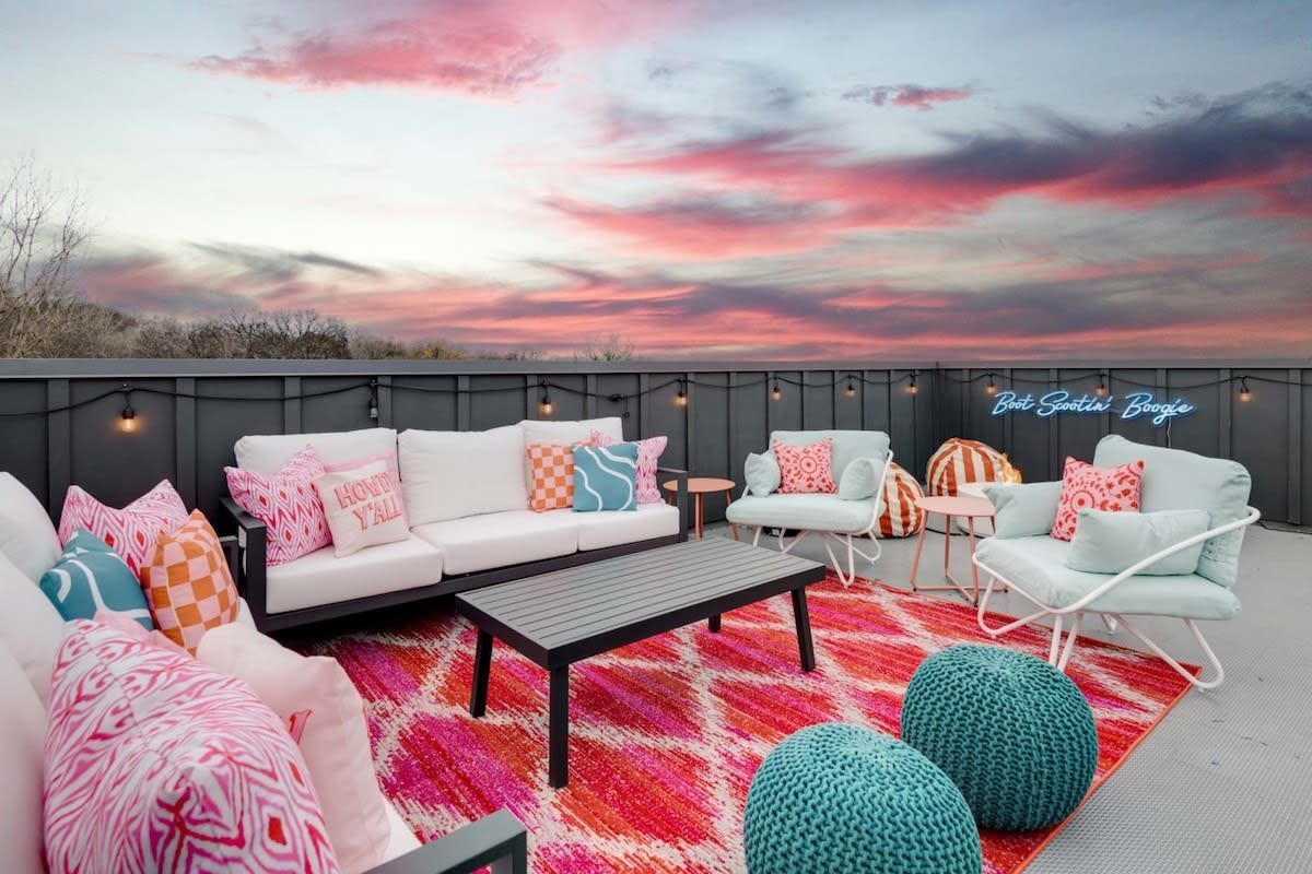 Outdoor rooftop lounge with white seating, pink patterned rug, and sunset sky with pink clouds