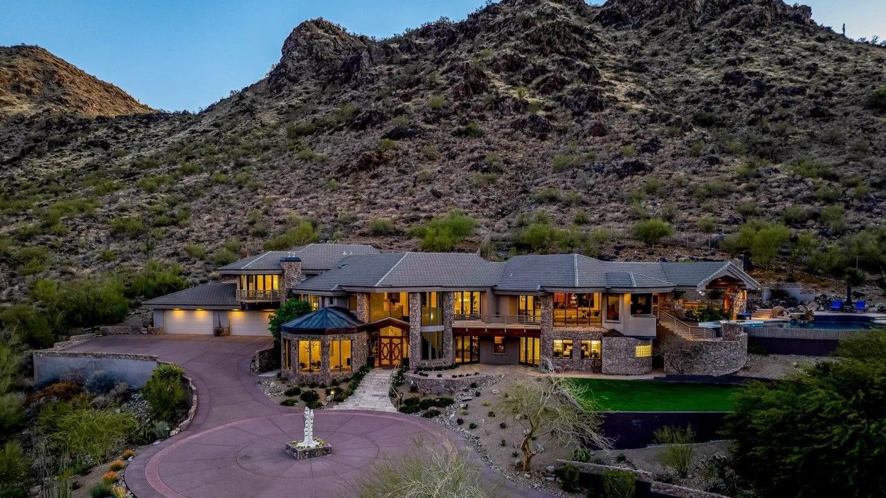 Luxurious house at the base of a rocky mountain. Long driveway, numerous windows, and manicured lawn.