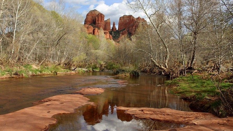 Red rock formations and bare trees frame a river, reflected in the water, under a cloudy sky.