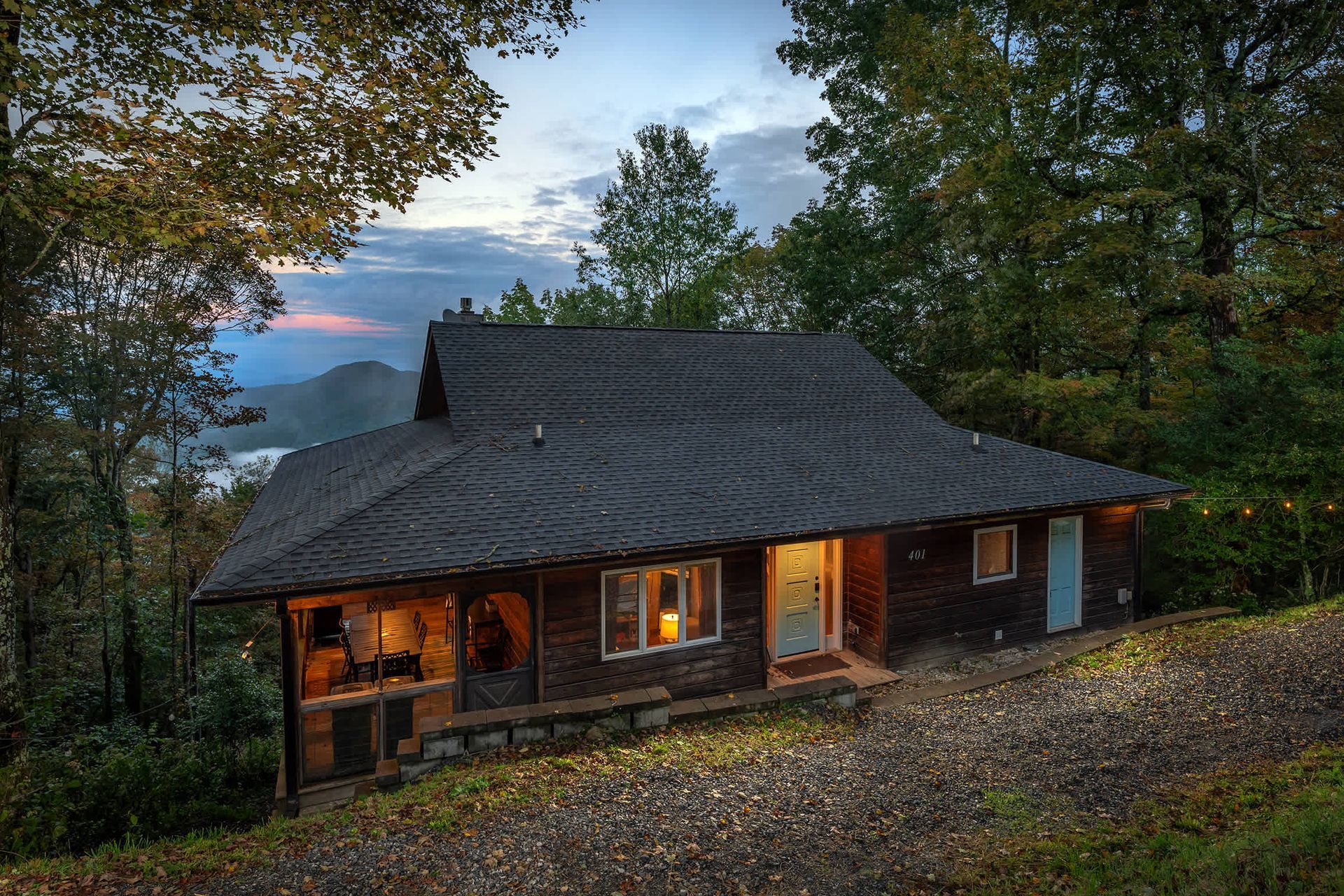 Cozy log cabin glowing at dusk on a wooded hillside with lit porch and windows