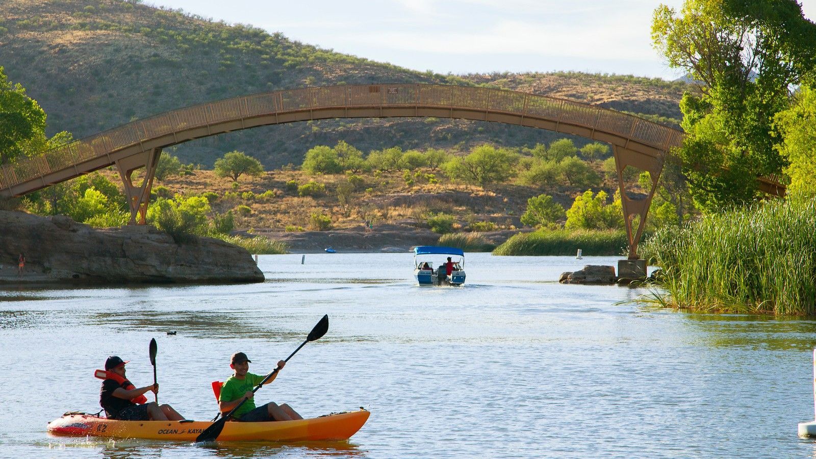 Two people kayak on a lake, under a wooden bridge; a boat with people passes by.