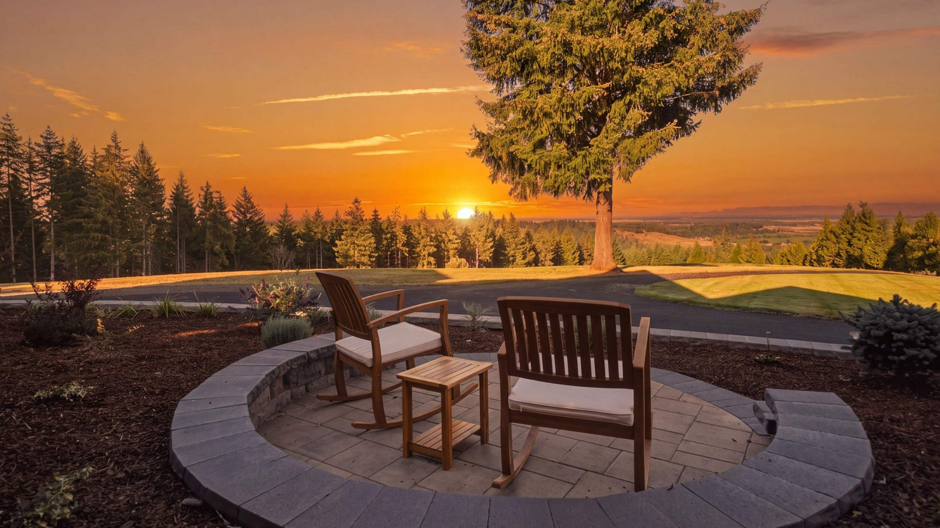 Two wooden chairs around a fire pit at sunset on a paved patio, with trees and a glowing horizon.