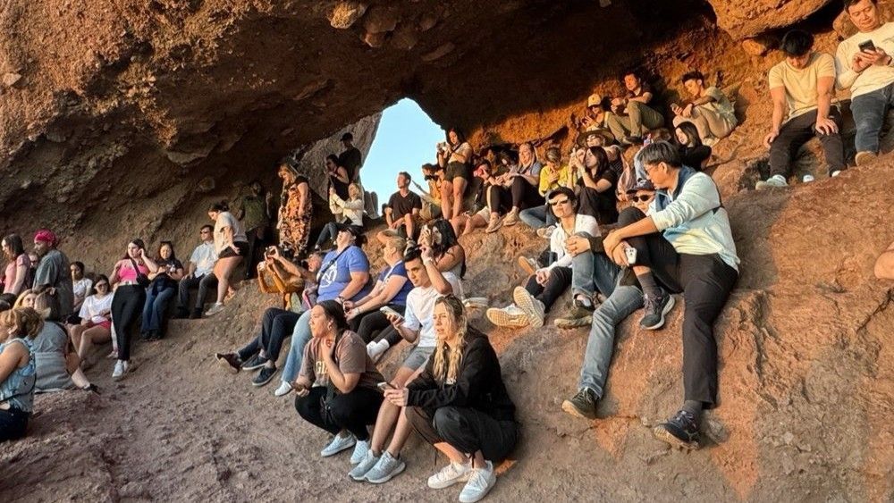 Large group of people gathered in a cave opening, watching something. Golden sunlight, rocky interior.