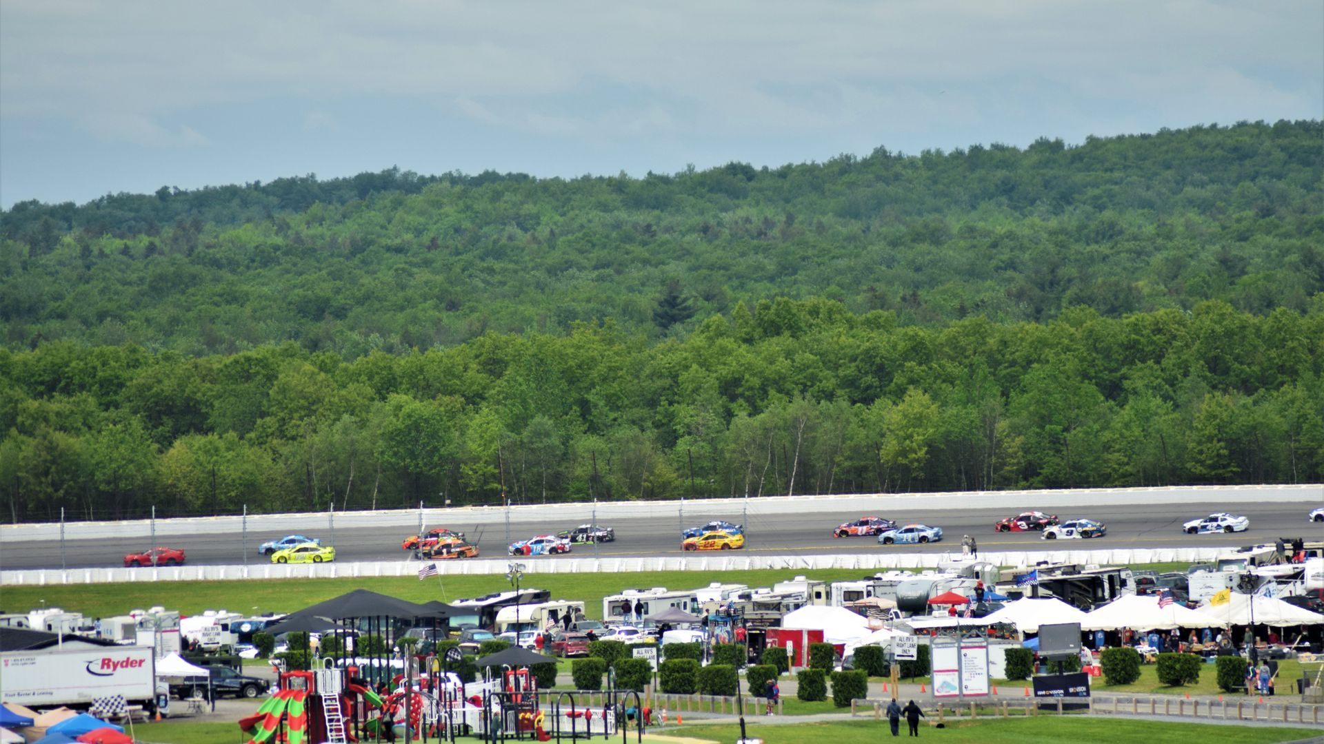 Race cars drive along a track with a lush, green forest in the background and an RV park in the foreground.