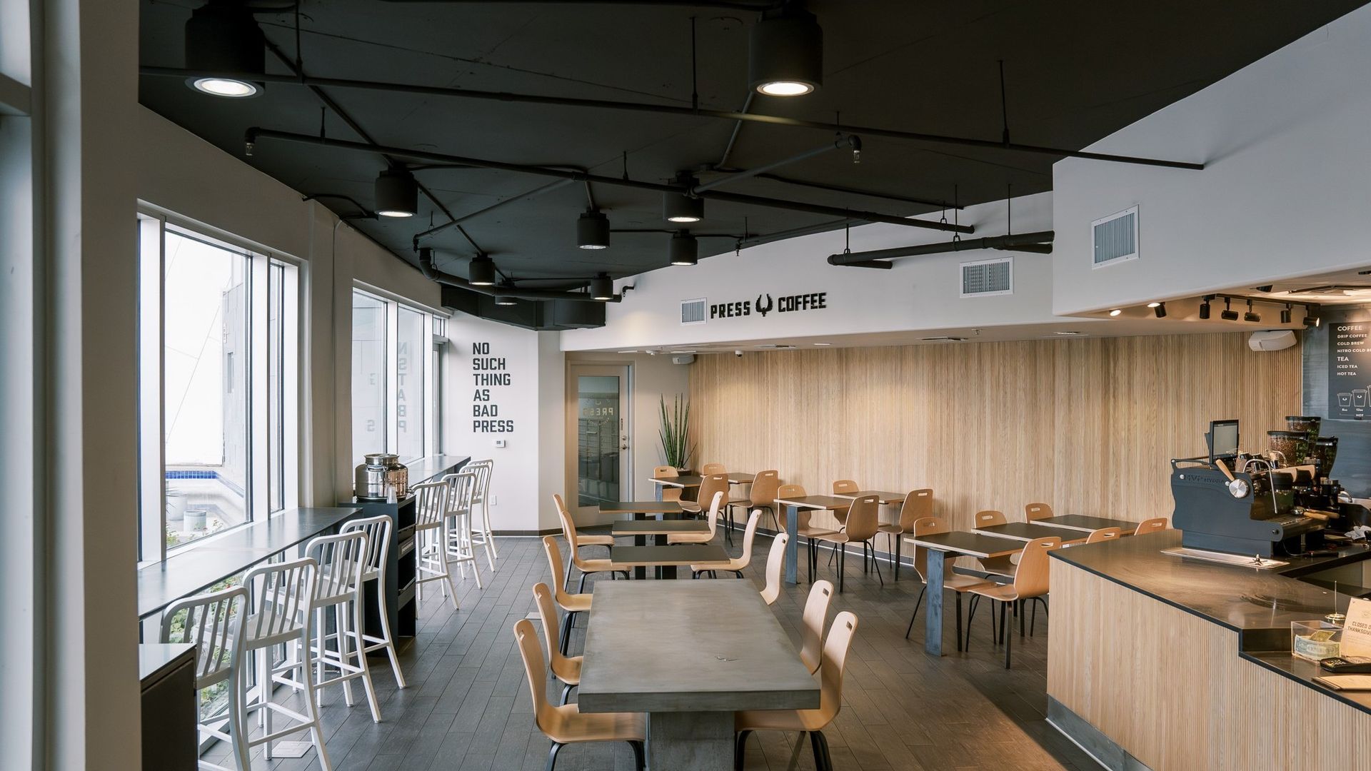 Interior of a cafe with concrete tables, wooden chairs, and large windows. The ceiling is black.