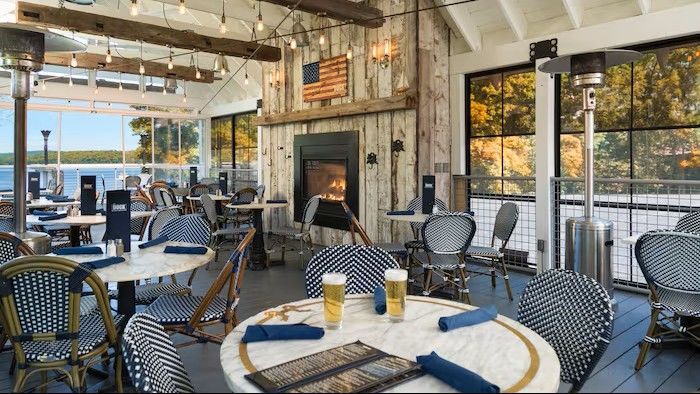 Outdoor restaurant seating with blue chairs, white tables, fireplace, and lake view.
