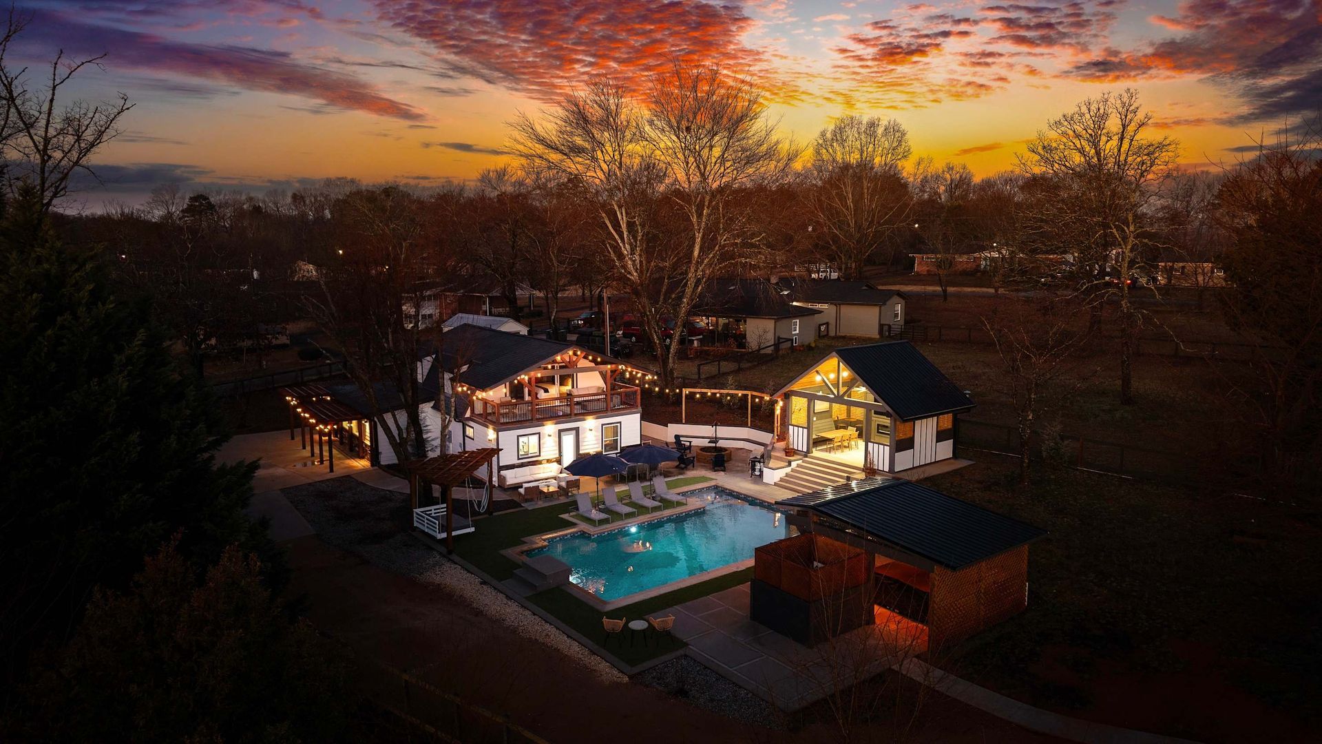 Sunset view of a lit house and pool surrounded by trees and a backyard deck