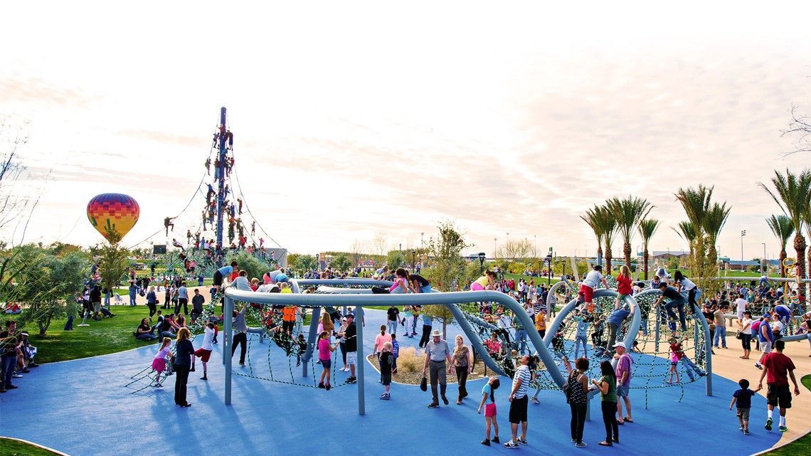 A large playground with many people. Blue ground, silver climbing structures, a hot air balloon, and palm trees.