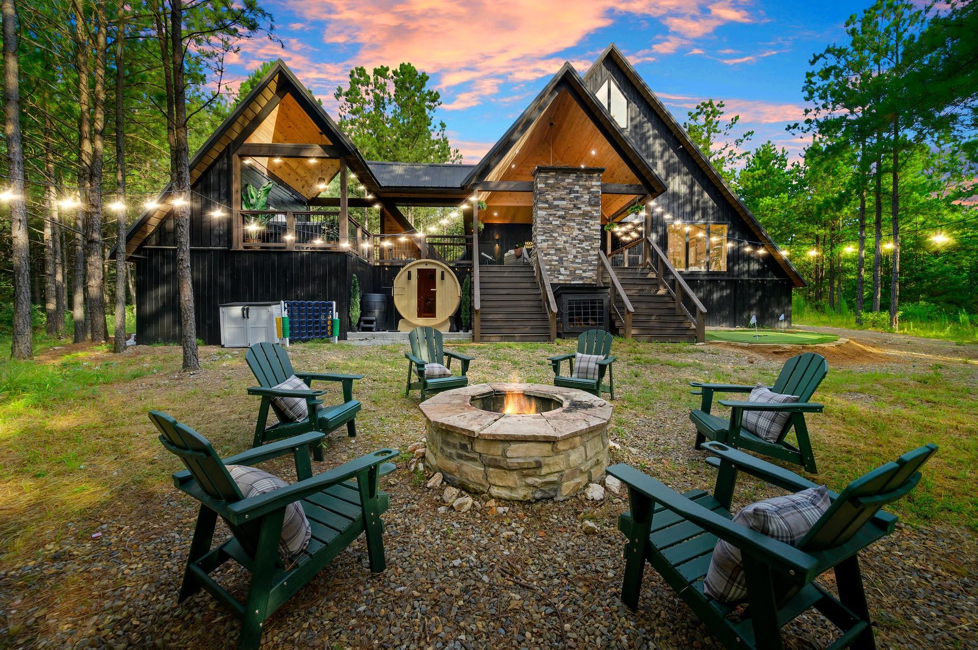 Rustic cabin with lit fire pit and green chairs in a wooded yard at dusk