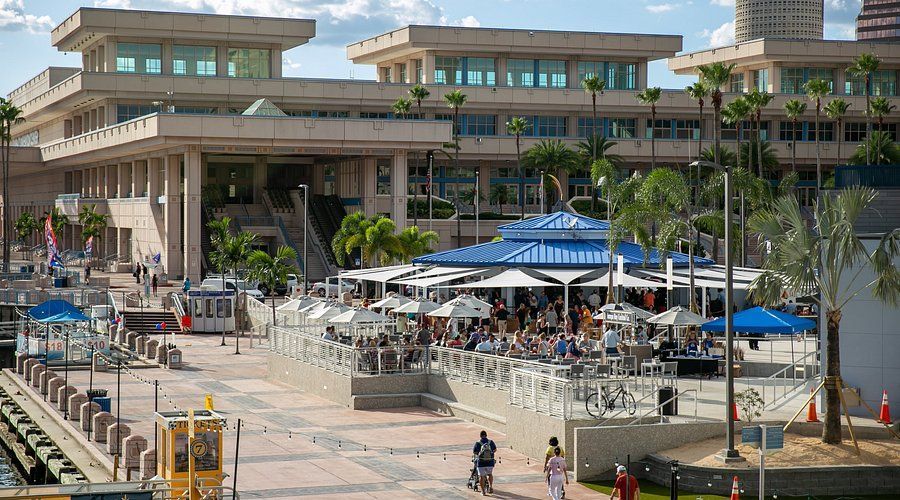 A waterfront restaurant with outdoor seating, with a large building behind it. People dine under umbrellas.