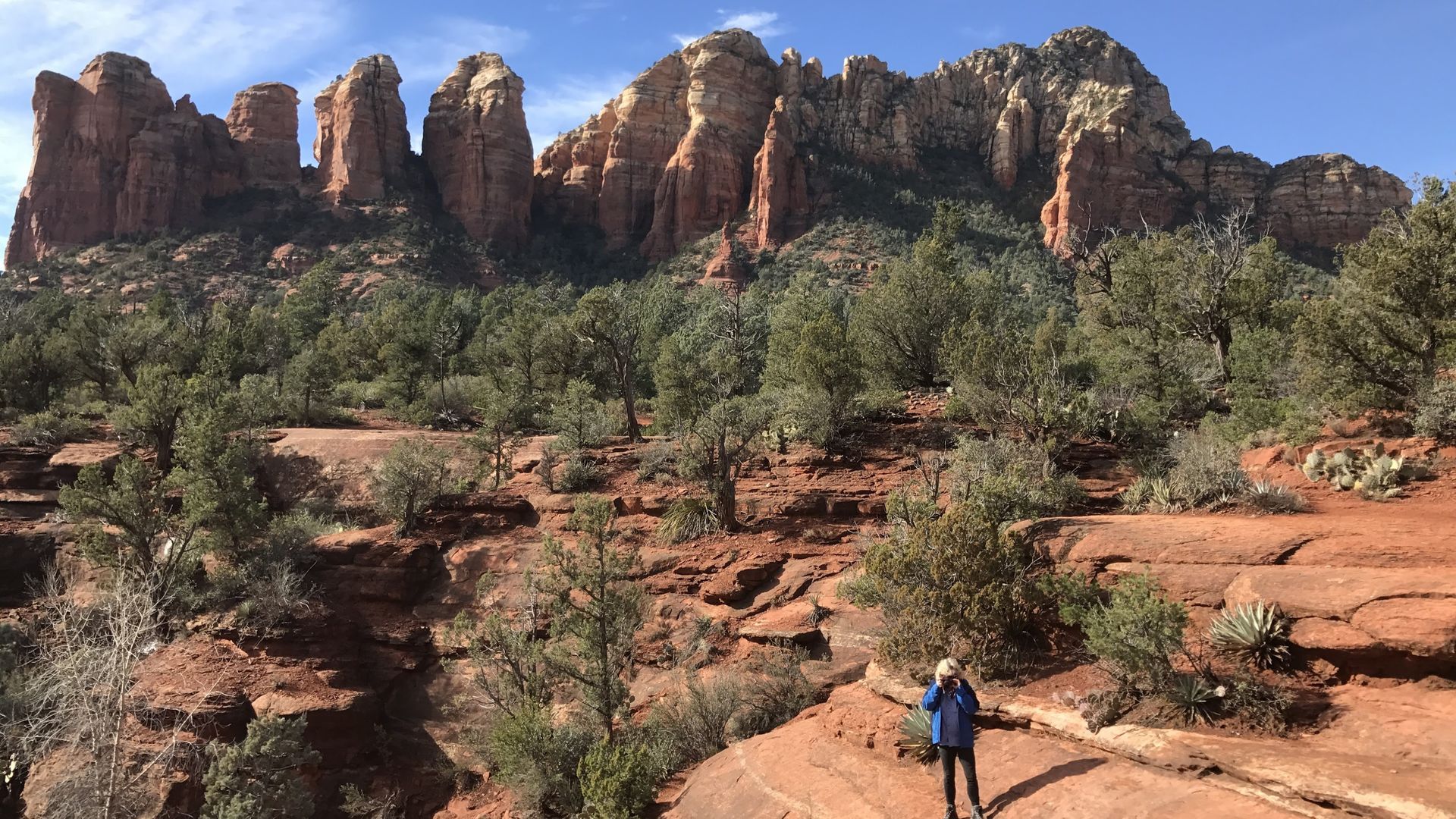 Person hiking on red rock trail with mountains in the background under a blue sky.