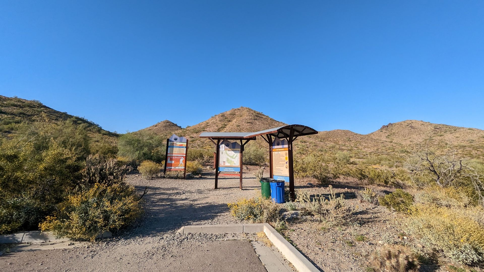 Trailhead with shelter and information boards; desert landscape and blue sky.