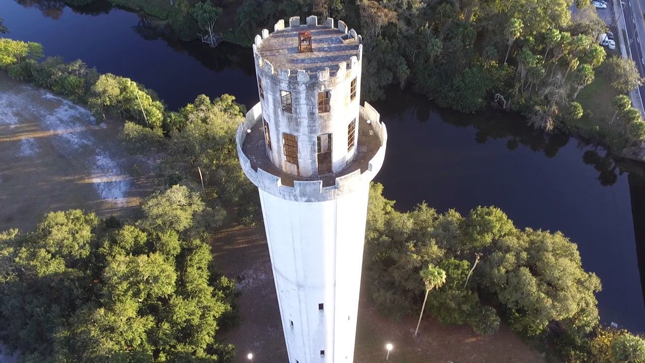 White water tower with castle-like top, standing near a river and trees.