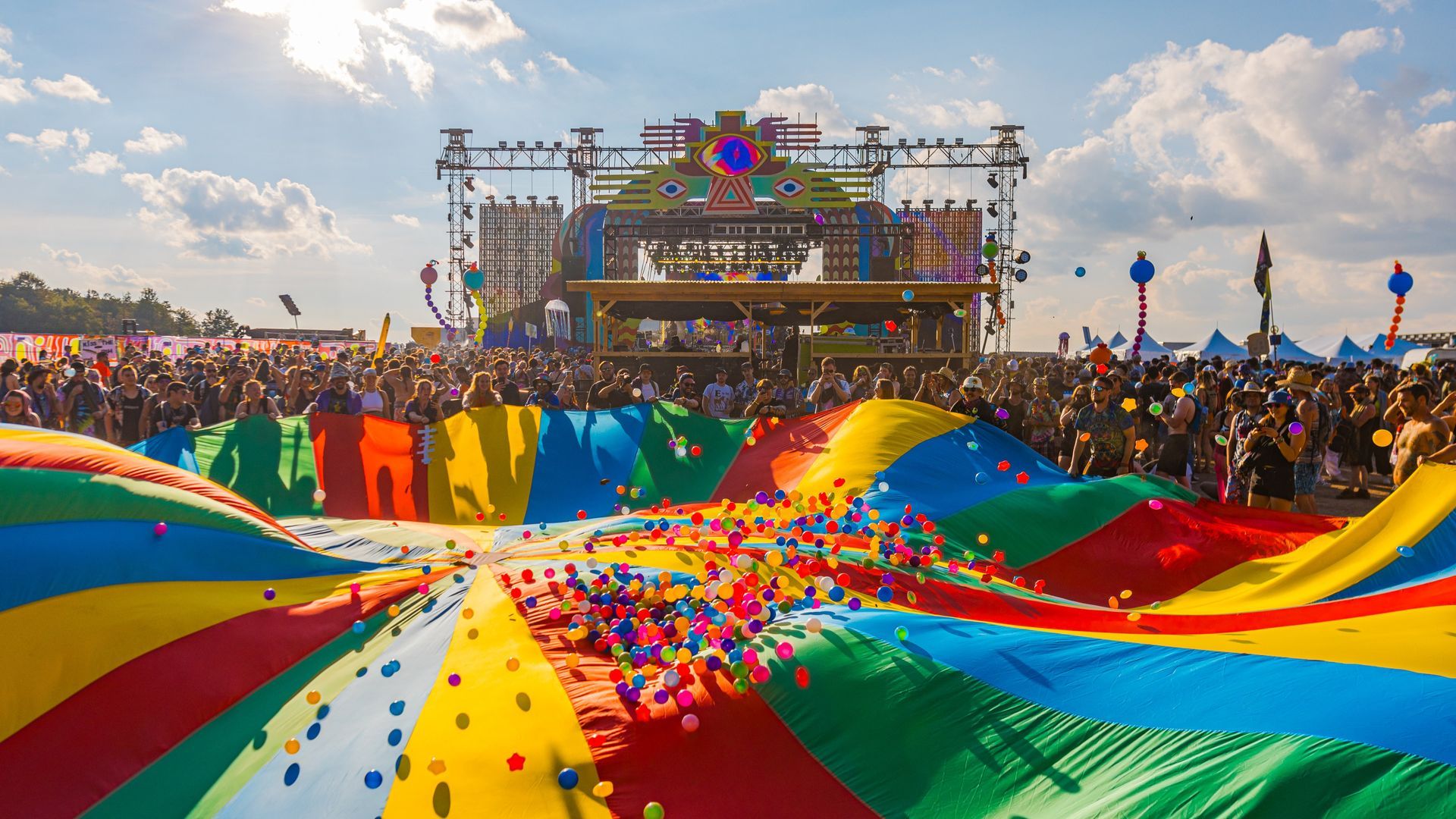 Large crowd at an outdoor music festival with a colorful fabric on the ground. Stage in the background.