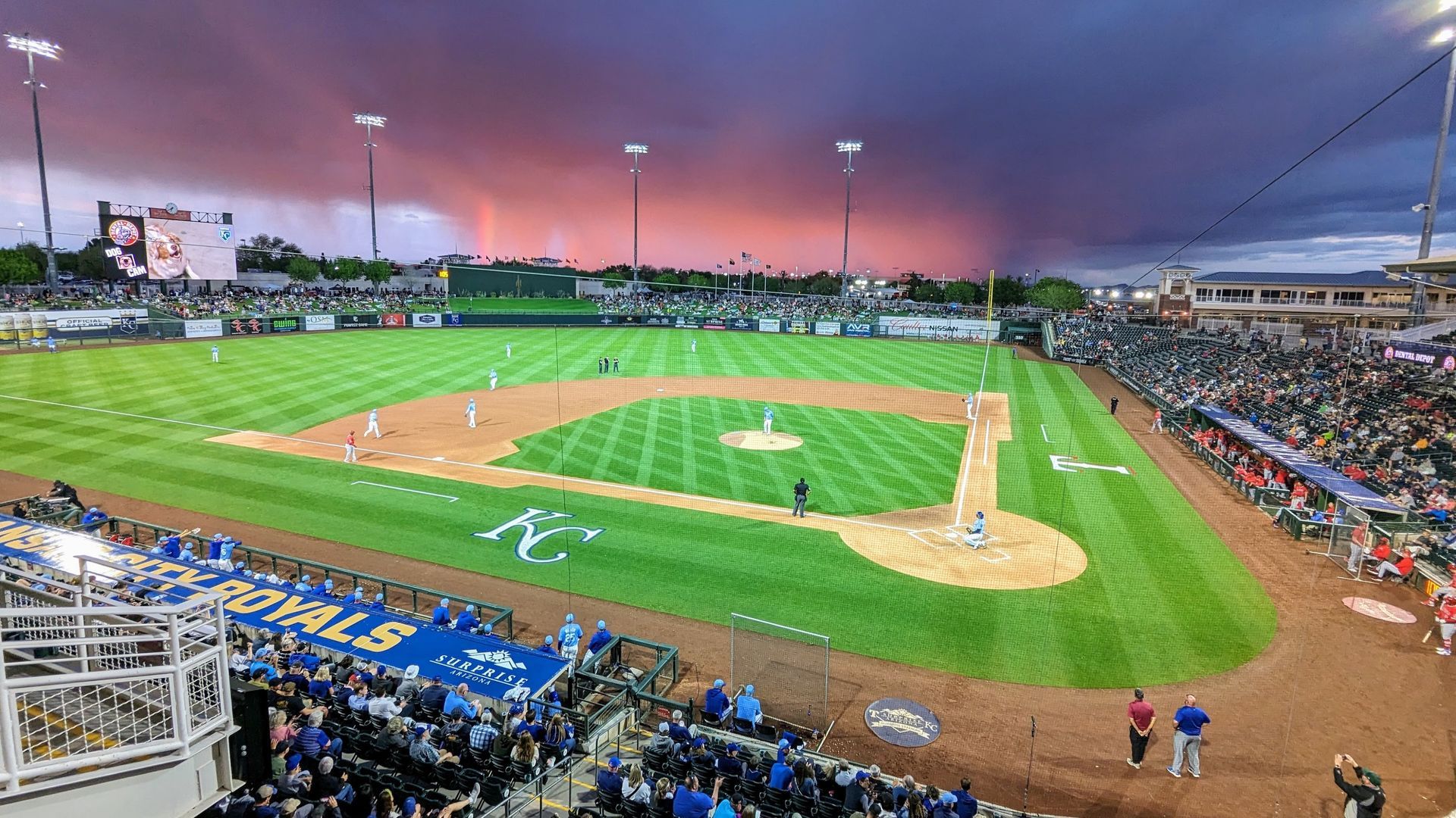 Baseball game in progress under a dramatic, colorful sunset. Stadium filled with fans.