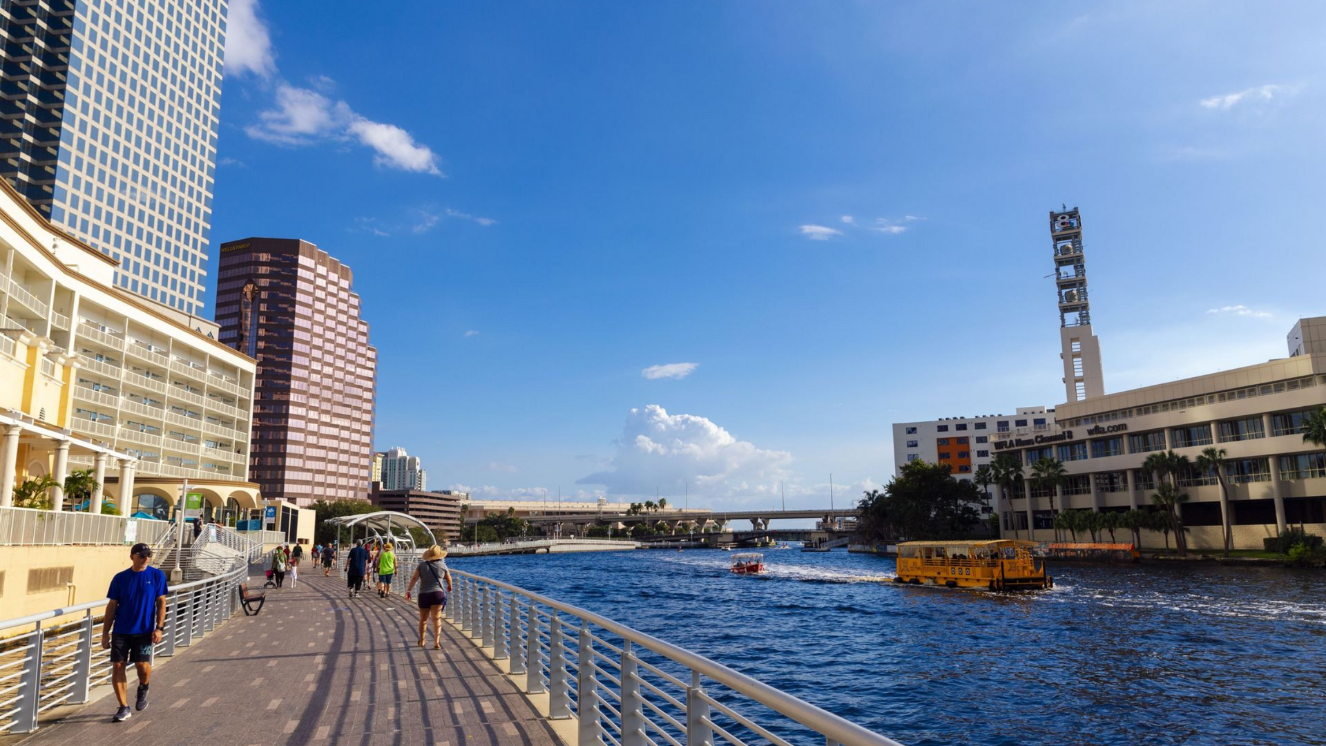 Waterfront scene with buildings, river, walkers, and a blue sky.