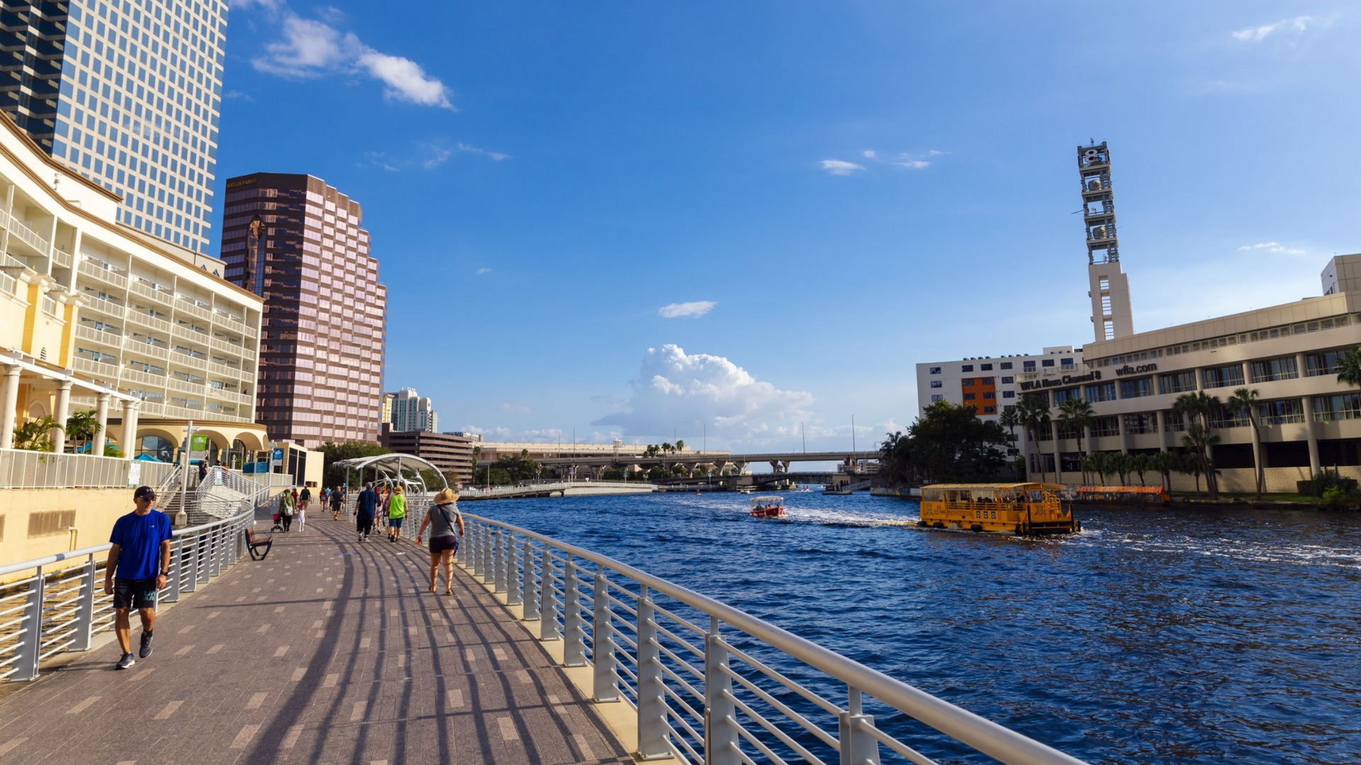 Waterfront walkway with people, buildings, bridge, and boat under a blue sky.