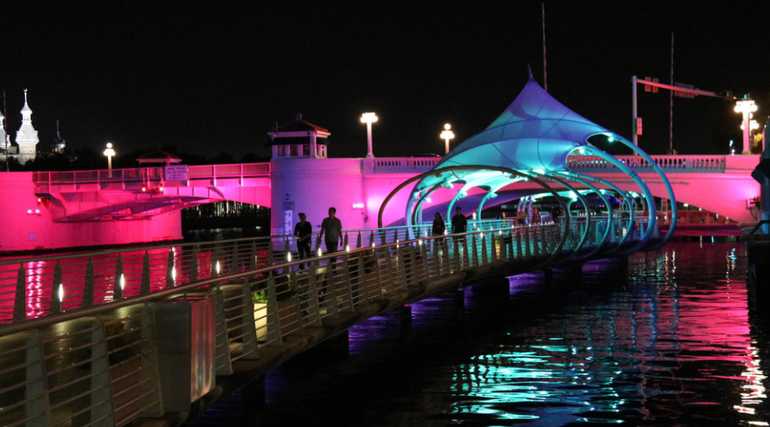 A brightly lit bridge over water at night. Two people walk towards the colorful canopy, with a castle in the distance.