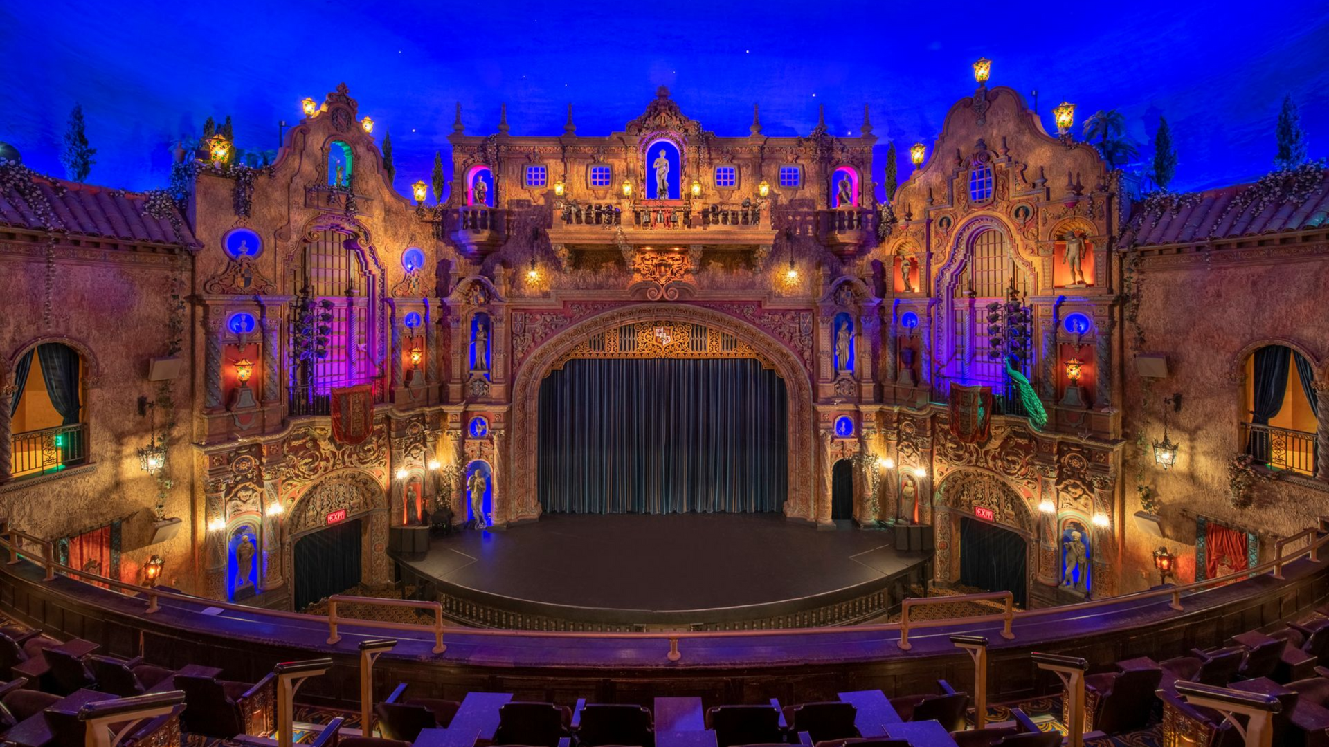 Ornate theater stage with gothic architecture, colorful lighting, and a closed stage curtain.