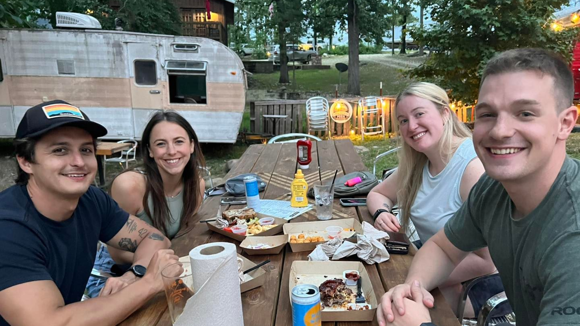 Four people smiling, seated at a picnic table with food, an old camper in the background.