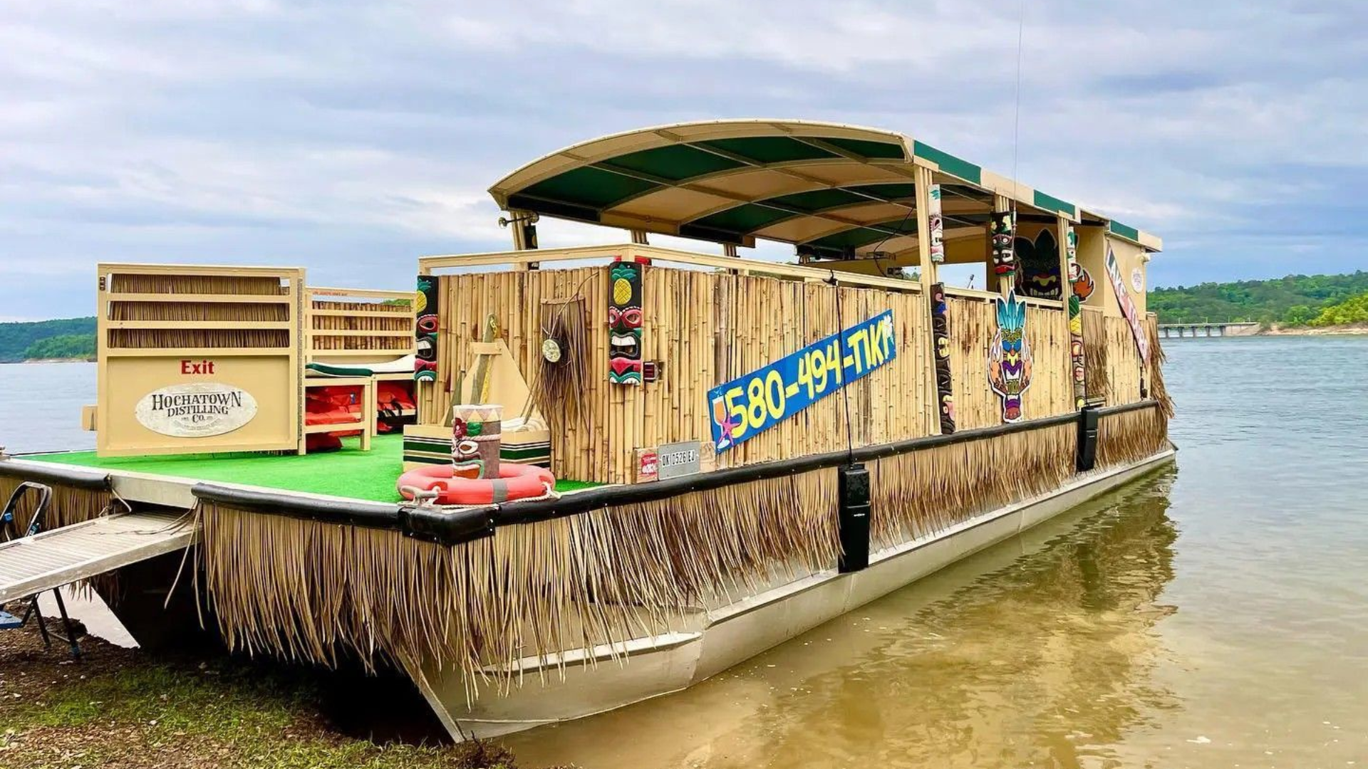 A themed pontoon boat with tiki decor docked on a lake shore under a cloudy sky.