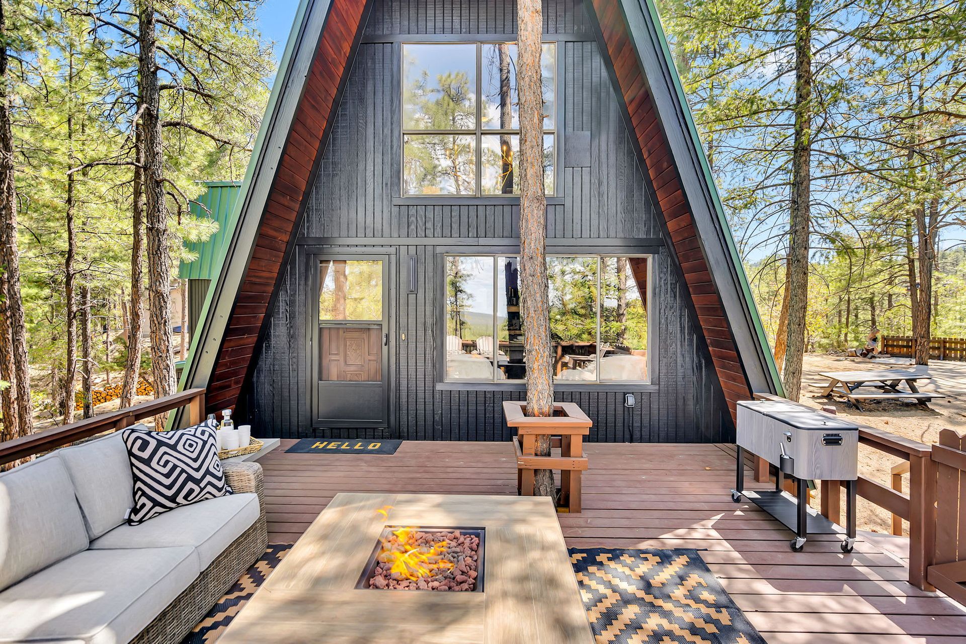 A-frame cabin deck with outdoor seating and fire pit, surrounded by tall pine trees.