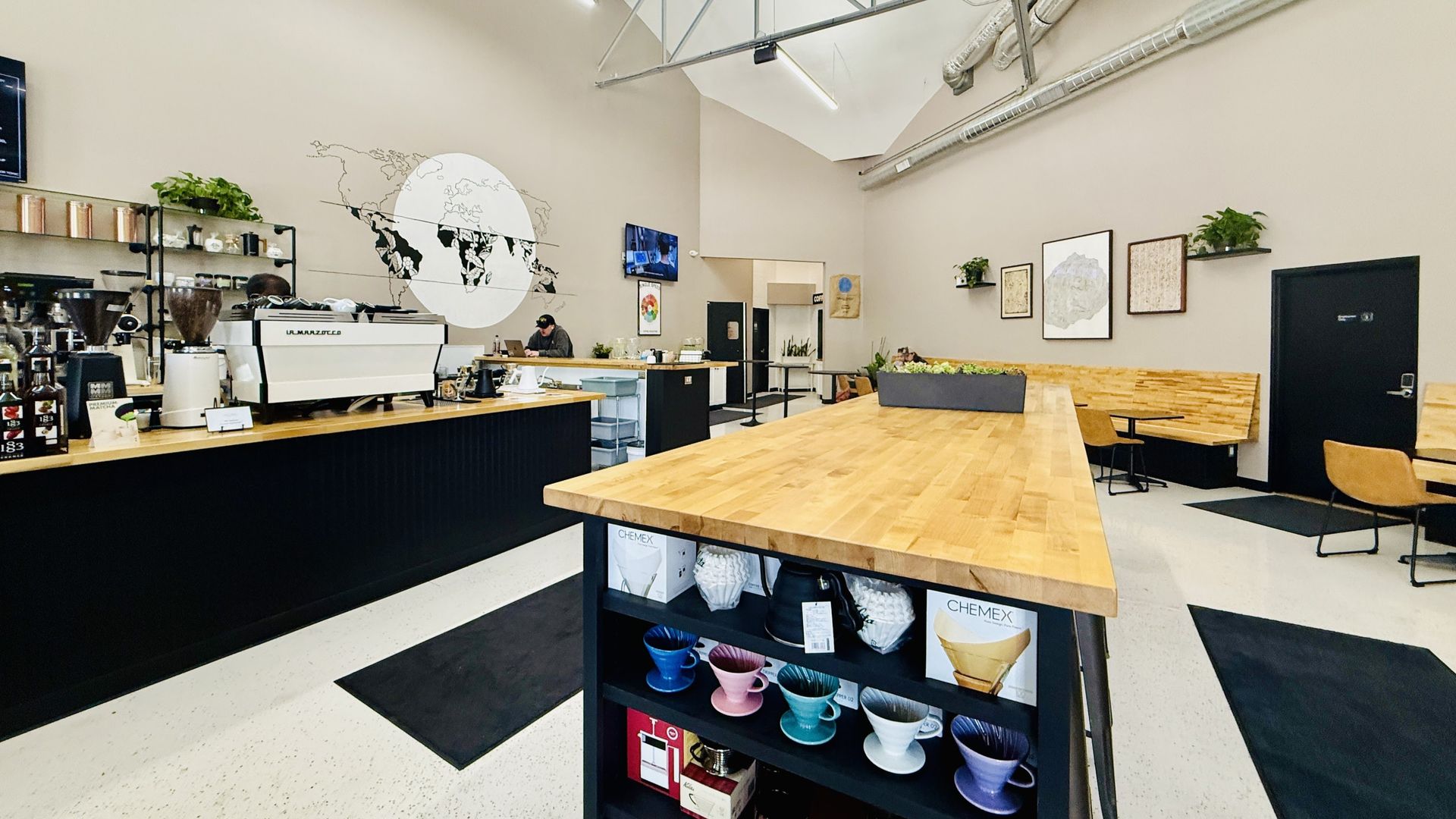 A modern coffee shop interior with a long wooden service counter, brewing equipment, and tables for seating.