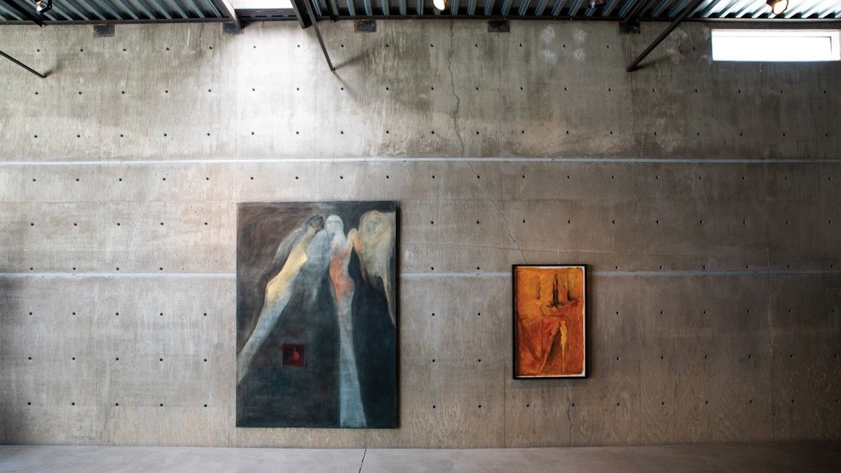 Two paintings hang on a concrete wall inside a building. Sunlight streams from above.
