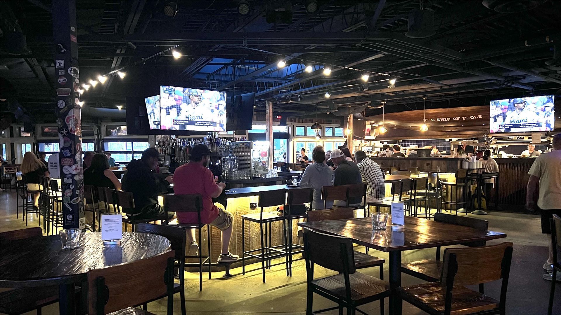 Interior of a bar, people sitting at tables and the bar, watching TVs. Dark setting with metal chairs.