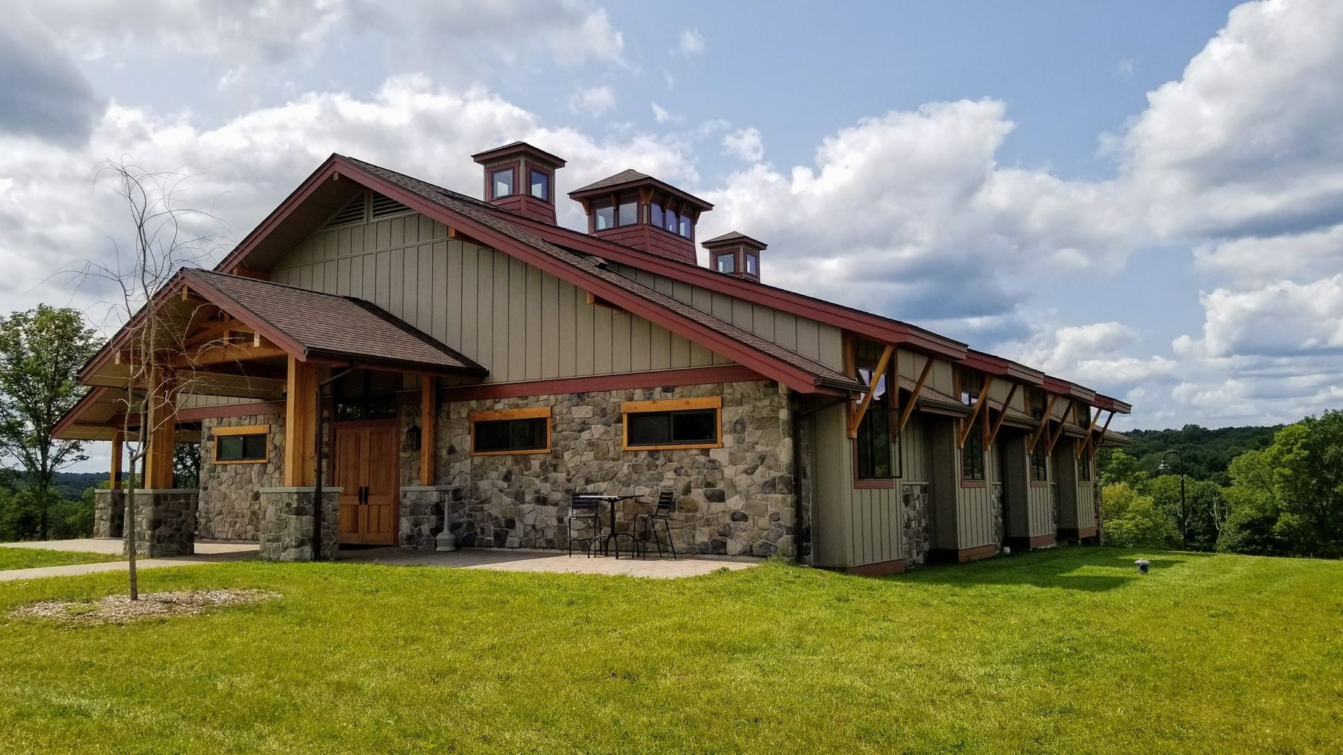 A stone and siding building with a reddish metal roof and three cupolas sits on a grassy hill under a cloudy blue sky.