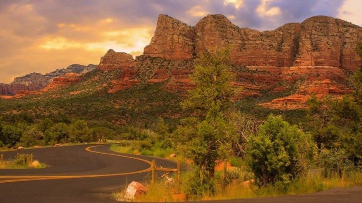 Winding road through a desert landscape; red rock mountains in the background under a cloudy sky.