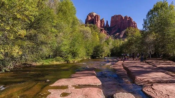 Red rock formations loom over a shallow river with red stone banks, flanked by green trees and foliage.