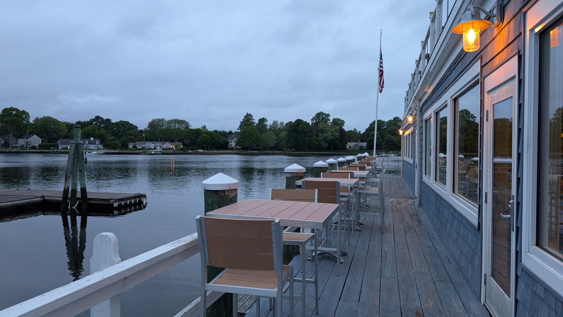 A waterside restaurant patio with tables and chairs, facing a river, under an overcast sky.