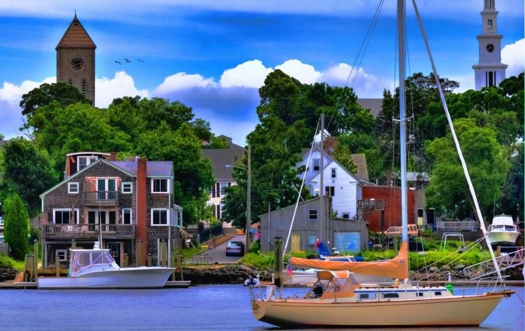Sailboat on water in front of a town with buildings, trees, and two tall towers under a cloudy sky.