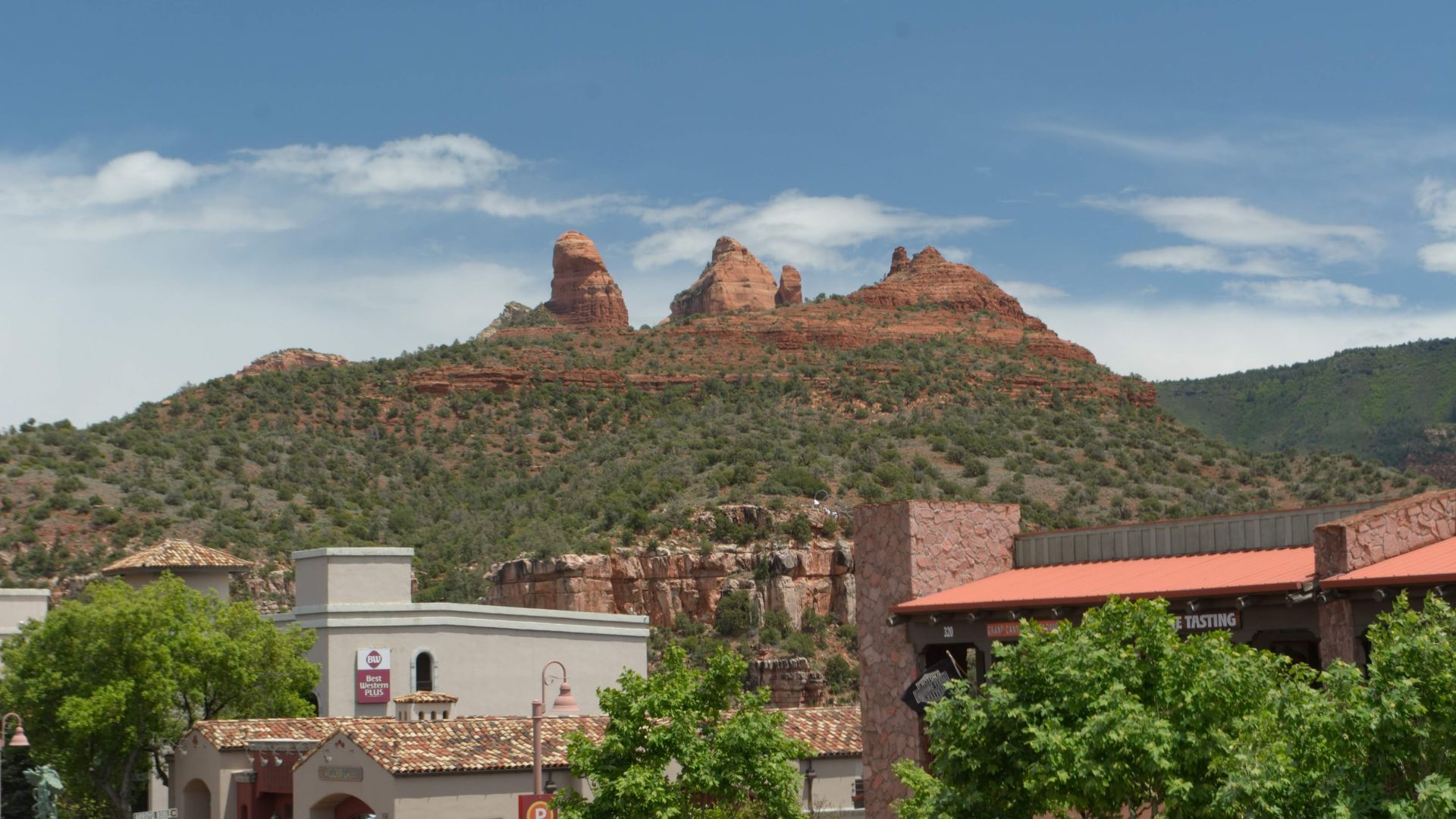 Red rock buttes rise above buildings with Spanish tile roofs and green trees, under a blue sky.
