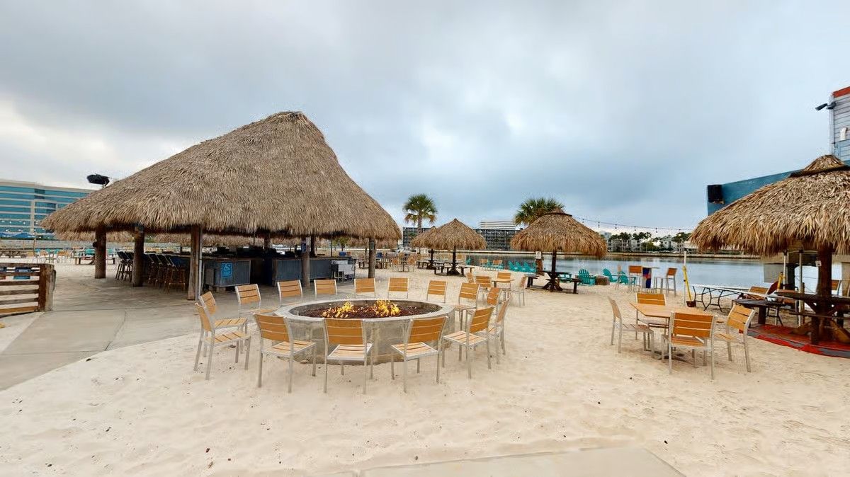 Outdoor beach bar with thatched roofs, sand, tables, fire pit, and water.