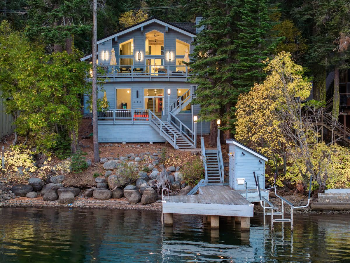 Blue lakeside cabin with lit windows, dock, and stairs among trees at dusk