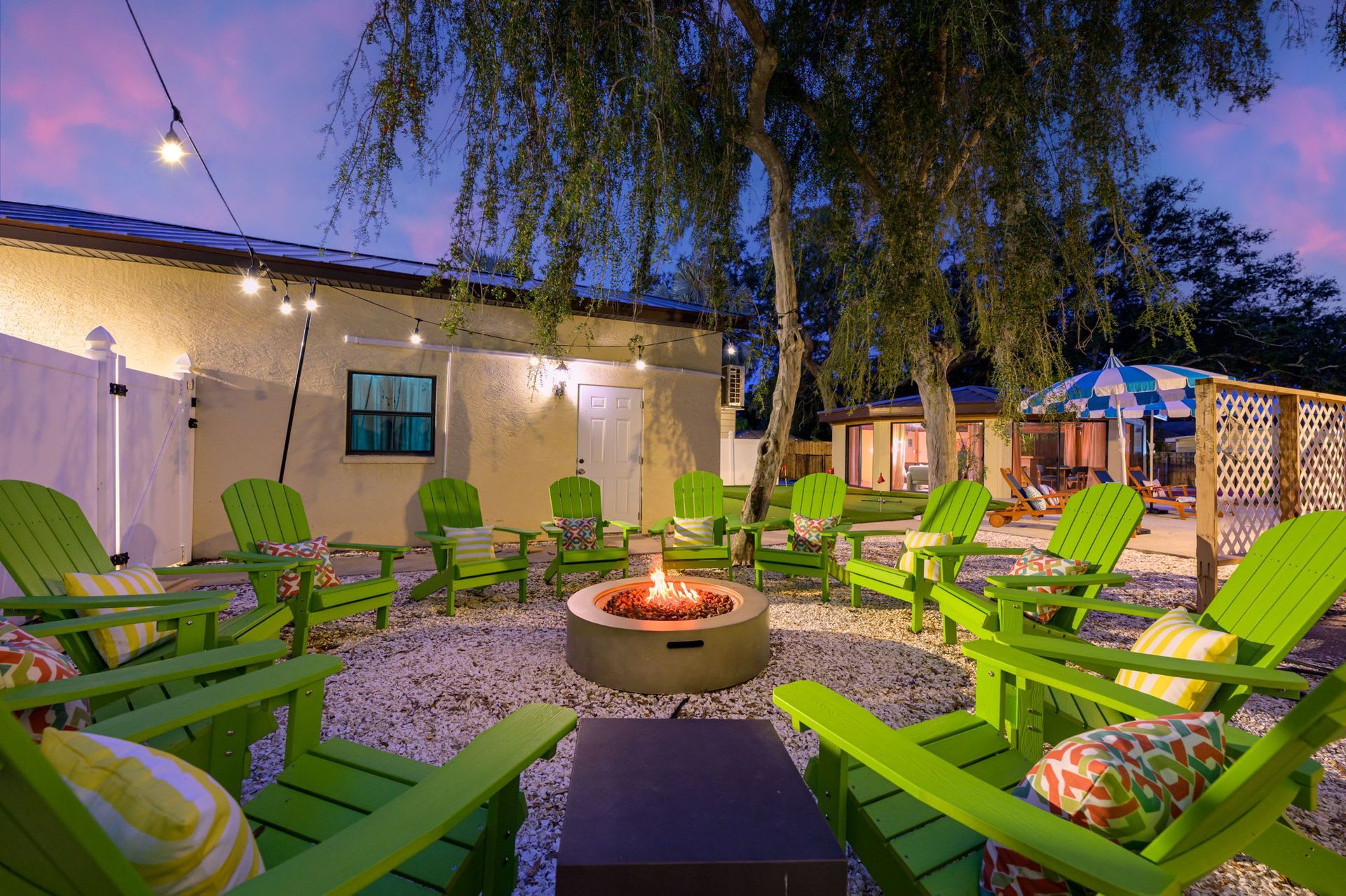 Outdoor patio with green lounge chairs around a fire pit at dusk, beside a lit building and trees