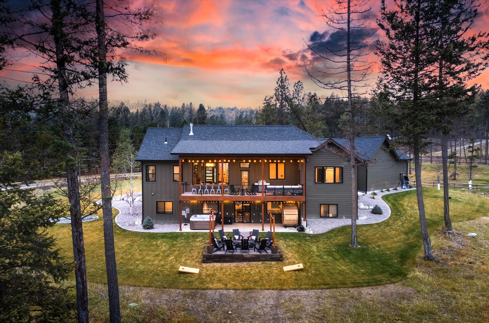 Two-story cabin lit warmly at dusk, centered in a wooded yard under a pink sunset sky.
