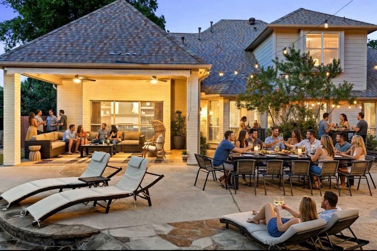 Backyard patio party at dusk with guests dining and lounging beside a lit modern house