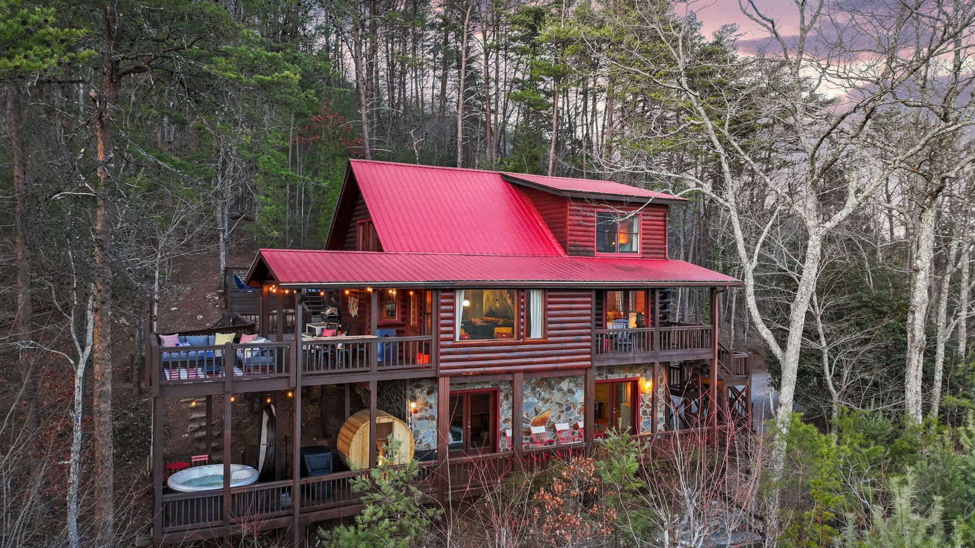 Red-roofed wooden cabin in a forest at dusk, with large windows and warm interior lights.