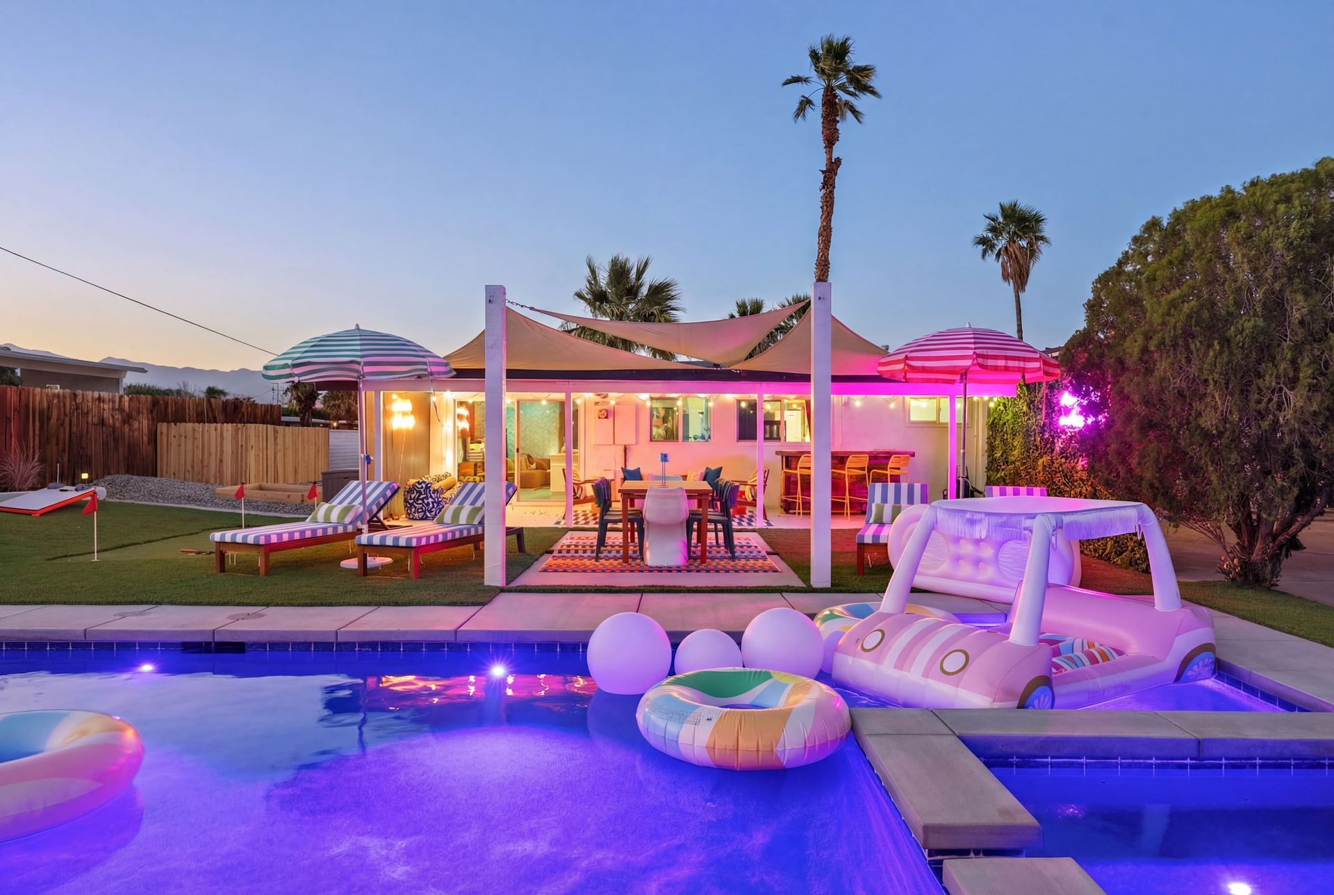 Lit backyard pool at dusk with a lit cabana, lounge chairs, and inflatable floats
