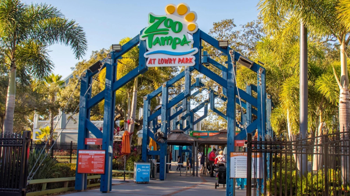 Zoo Tampa entrance with blue archway and sign, people entering, trees in background.