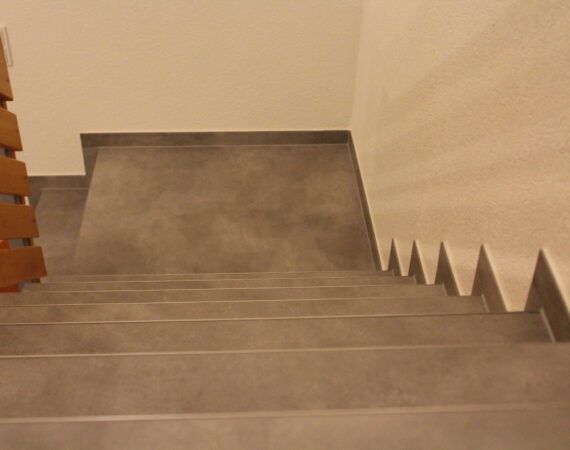Blick hinunter auf eine grau geflieste Treppe, rechts eine weiße Wand, links ein Holzgeländer.