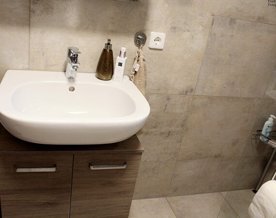 Waschbecken im Badezimmer mit Armatur, Schrank, neutral gefliesten Wänden und Toilettenartikeln.