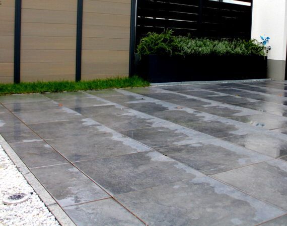 Grau geflieste Terrasse mit nasser Oberfläche, im Hintergrund Gras und eine Holzwand.