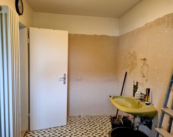 Badezimmerrenovierung im Gange: freiliegende Wand, gelbes Waschbecken, offene Tür und Schachbrettmusterboden.
